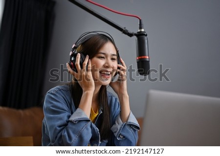 Attractive young Asian female wearing headphones speaking on her microphone, recording her podcast contents, running her online radio in studio at home. 