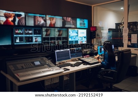 Middle aged beautiful woman working in a broadcast control room on a tv station. Entertainment news with footage equipment concept. Focus on a equipment on the table. Royalty-Free Stock Photo #2192152419