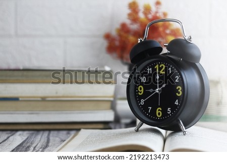 Black alarm clock, with an open book on a wooden table and a white textured wall background, educational concept.