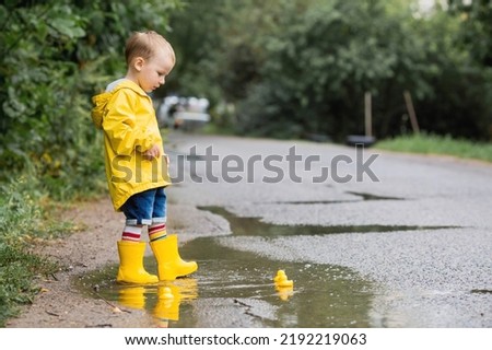 A small child in rainbow socks, yellow rubber boots and a jacket jumps through puddles and plays with yellow rubber ducks. A picture of summer and autumn holidays. A child in the rain.