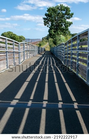 Public footbridge in stark shadowed sunlight. Long stripes of black shadow follow the contours of the bridge. Used by cyclists, walkers and hikers. strong picture of contrast between light and shadow.