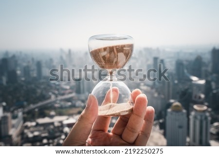 Hand holding a Hourglass with cityscape on panoramic skyline and buildings in the morning background with sun light. The concept of modern life, business, time, management and city life.
