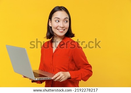 Excited smiling happy fancy vivid young woman of Asian ethnicity 20s years old wears orange shirt hold use work on laptop pc computer looking back isolated on plain yellow background studio portrait