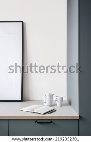detail of interior design with ceramic cup and teapot, opened blank notepad with pen and white poster on wall in modern apartment, cropped shot
