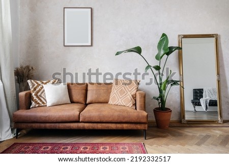 Modern design of living room with brown eco leather couch, soft cushions, mirror with golden frame, copy space picture frame on wall and houseplant in pot Royalty-Free Stock Photo #2192332517