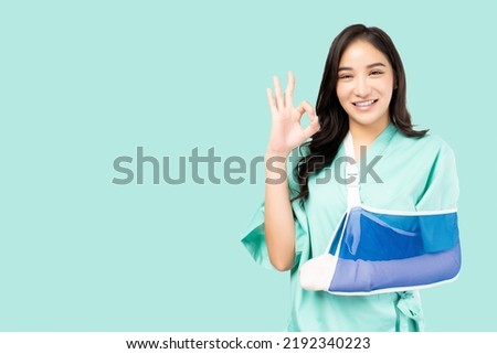 Beautiful young asian woman wearing patient gown and soft splint and wrapped in bandages on arm showing okay sign on green background Patient Insurance Health care and Personal accident concept