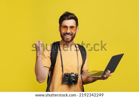 Online tour booking concept. Overjoyed man with backpack and camera using laptop computer to book his journey over internet, shaking clenched fist over yellow studio background