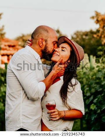 Portrait of a smiling  happy couple kissing    in a Vineyard toasting wine. Beautiful  brunette woman and bearded muscular man spending time together during grape harvest.