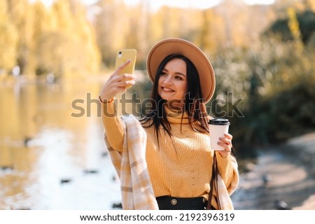Portrait of a happy smiling young woman millennials in a hat laughing talking using a mobile phone taking a selfie walking alone in an autumn park in nature in autumn. Selective focus