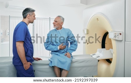 Radiologic technologist listening to senior patient's complaints sitting on CT scanner bed. Computed Tomography with contrast Royalty-Free Stock Photo #2192397039