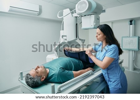 Female technician preparing patient for body X-ray in radiographic imaging room, putting for him radiation protection apron Royalty-Free Stock Photo #2192397043
