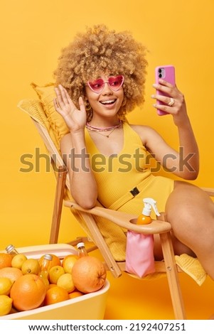 Positive curly woman wears heart shaped pink sunglasses and bathing suit waves hello at smartphone camera has video talk with best friend shares impressions of spending summer vacation at resort