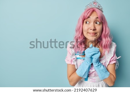 Horizontal shot of pink haired housewife keeps hands together wears crown white dress and rubber gloves concentrated aside isolated over blue studio background empty space for your promotion