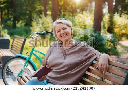 smiling modern old lady relaxing city park. Pensive senior gray haired woman casual sitting bench outdoors reading book. cycling forest park, bicycle, healthy active lifestyle after 50-60 years Royalty-Free Stock Photo #2192411889