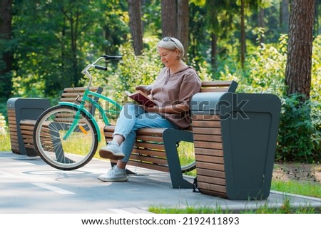 Dreamy modern old lady relaxing city park. Pensive senior gray haired woman casual sitting wooden bench outdoors reading book. cycling forest park, bicycle, healthy active lifestyle after 50-60 years Royalty-Free Stock Photo #2192411893