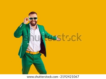 Happy cheerful handsome young man in stylish green suit and sunglasses isolated on yellow color background smiles, invites you to St Patrick's Day party and shows something on empty blank copy space