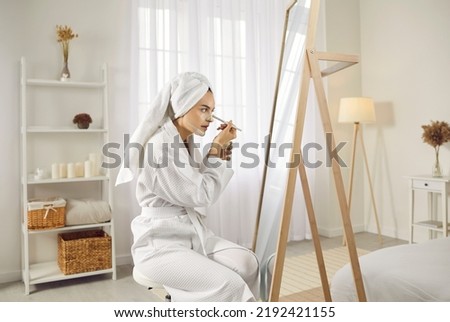 Beautiful young woman in a white dressing gown and bath towel turban on her hair sitting in front of a big mirror in the bedroom, looking at her reflection and applying facial beauty mask on her Royalty-Free Stock Photo #2192421155