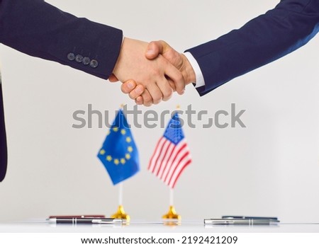 Two diplomats from USA and Europe shake hands at negotiation table with flags of US and EU in background. Politicians work toward developing foreign trade and establishing good friendly relationship