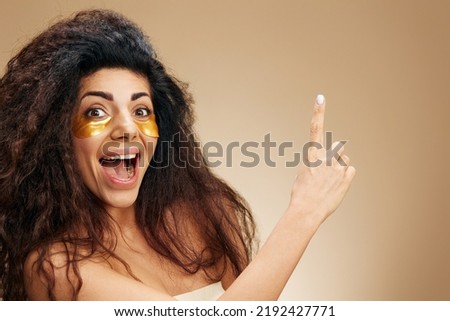 Amazed curly pretty Latin female with gold hydrogel patches under eye point finger up open mouth posing isolated over pastel beige background. Cosmetic product ad Natural beauty concept Copy space