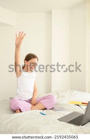 10-year-old schoolgirl with raised hand sitting on bedroom, child looking at laptop. Online class, home education, training, knowledge, comfortable online school concept. Caucasian girl. Day light