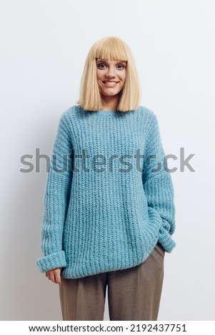 vertical photo of a beautiful, calm woman in a blue sweater standing on a light background with her hand in her pants pocket