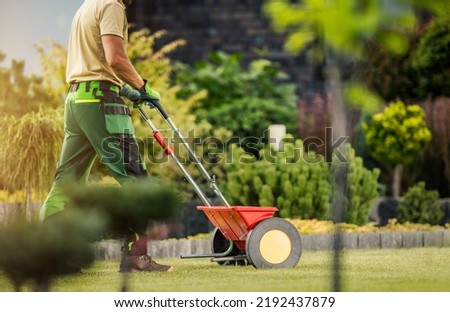 Caucasian Professional Gardener with Push Spreader Fertilizing Residential Lawn For a Good Health and Appearance of the Grass. Garden Maintenance. Royalty-Free Stock Photo #2192437879
