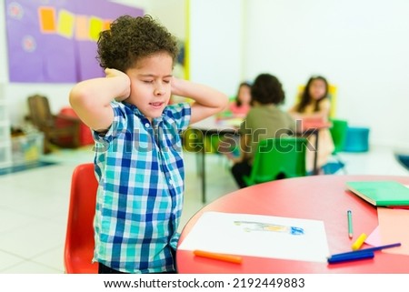 Upset autistic little boy covering his ears and feeling distressed and overwhelmed by the loud noises in preschool  Royalty-Free Stock Photo #2192449883