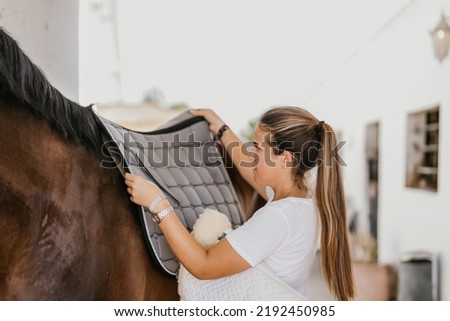 young woman placing a blanket over the back of a horse to fit a saddle Royalty-Free Stock Photo #2192450985