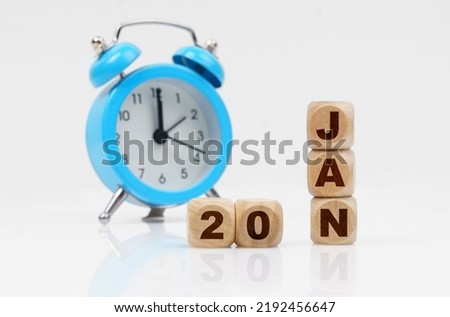 20st of January. Wooden cube calendar for January 20, next to a blue alarm clock. Objects on a reflective white surface