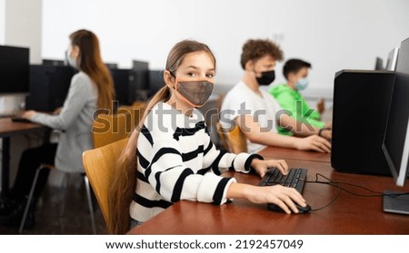 Interested teen girl in protective face mask studying with classmates in school computer class. Necessary pandemic precautions..