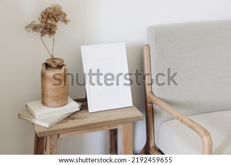 Empty vertical picture frame mockup. Ceramic vase with dry hydrangea flowers on pile of old books. Midcentury linen sofa. White wall background. Scandinavian interior, living room design.
