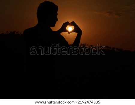 Silhouette photo of guy making heart gesture and orange sun in between.