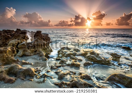 This early morning sunrise picture has rocks in the foreground with sun beams of light radiating outwards in the blue sky.  This picture was taken on the famous Jupiter Island.
