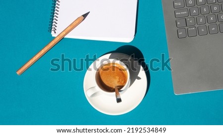 Blank page of a notebook next to a laptop and a white cup with coffee on a blue background