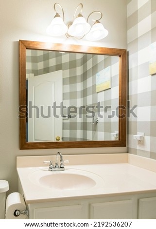 Vertical bathroom interior with checkered wallpaper on the right. There is a toilet with potted plant and picture frame near the framed mirror on the right above the vanity sink with white drawers.