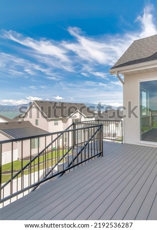 Vertical Whispy white clouds Deck of a house with stairs, wood flooring, and metal railings. There is a sliding glass door near the stairs and windows on the right with a view of the neighborhood