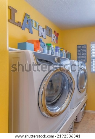 Vertical Laundry room interior with decorations on the yellow wall. There are front load laundry appliances on top of a base platform with baskets at the bottom across the wall mounted hooks