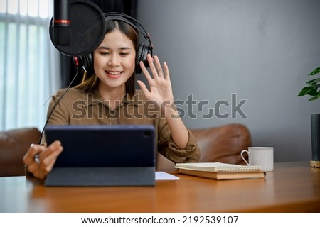 Beautiful young Asian female radio host wearing headphones, speaking on her microphone, running her radio show or podcasting at home studio. Work from home concept