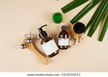 A mock-up of a brown cosmetics bottle with dispenser and white label, round green cream container, bottle with pipette, face brush, face massage roller on beige colored background lined up, top view