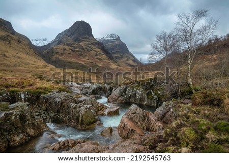 Beautiful landscape image of vibrant River Coe flowing beneath snowcapped mountains in Scottish Highlands 