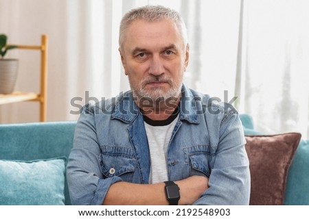 Angry senior man with gray hair and crossed arms looking at camera, sitting on sofa at home  Royalty-Free Stock Photo #2192548903