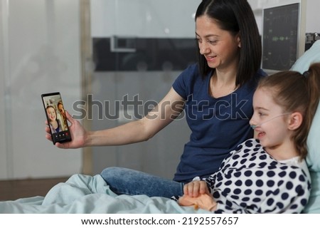 Mother and sick daughter talking on smartphone virtual videocall with best friend while resting on patient bed inside hospital pediatrics ward room. Ill little kid talking on online phone call. Royalty-Free Stock Photo #2192557657