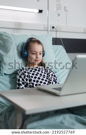 Sick girl with headphones watching funny cartoons on laptop while sitting on patient bed inside hospital pediatrics ward room. Ill kid under treatment enjoying internet video content inside clinic.