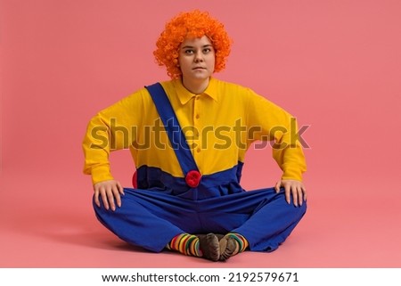a cheerful clown in a wig and a yellow-blue suit sits cross-legged on a colored background