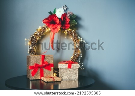 Few glittering golden gift boxes with velvet red ribbon and elegant Christmas flower wreath on the black table on gray wall background. Minimalist festive home decor. Scandinavian style. Copy space.