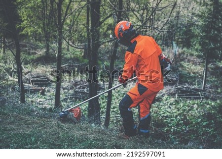 Man holding a brushcutter cut grass and brush. Lumberjack at work wears orange personal protective equipment. Gardener working outdoor in the forest. Security, occupation, forestry, worker, concept Royalty-Free Stock Photo #2192597091