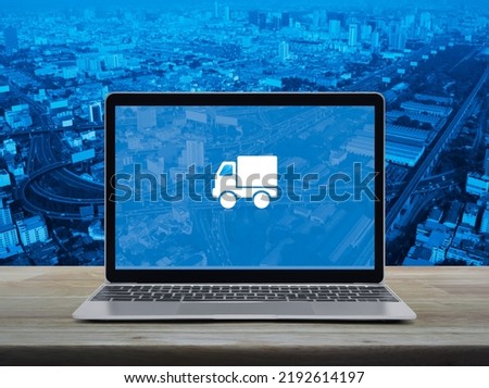 Delivery truck flat icon on modern laptop computer screen on wooden table over city tower, street, expressway and skyscraper, Business transportation online service concept