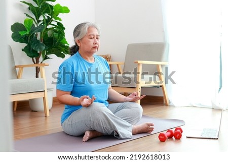 Asian elderly woman sitting at home exercising, doing exercises according to online fitness trainers. through a video call on a laptop. Social distancing. meditate doing yoga