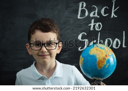 Cute caucasian schoolboy wearing glasses and holding the Globe against the black school board in the classroom at elementary school. Back to school concept
