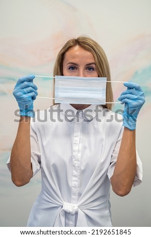 A beautiful young woman wearing a medical mask on a light background. The nurse puts a protective mask on her face. Measures to prevent coronavirus